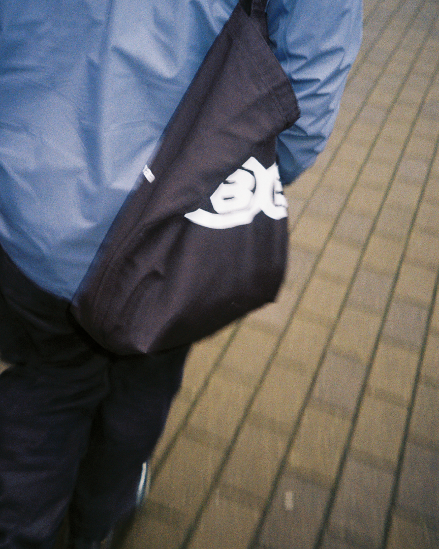 A figure wears a blue jacket and a branded fabric bag